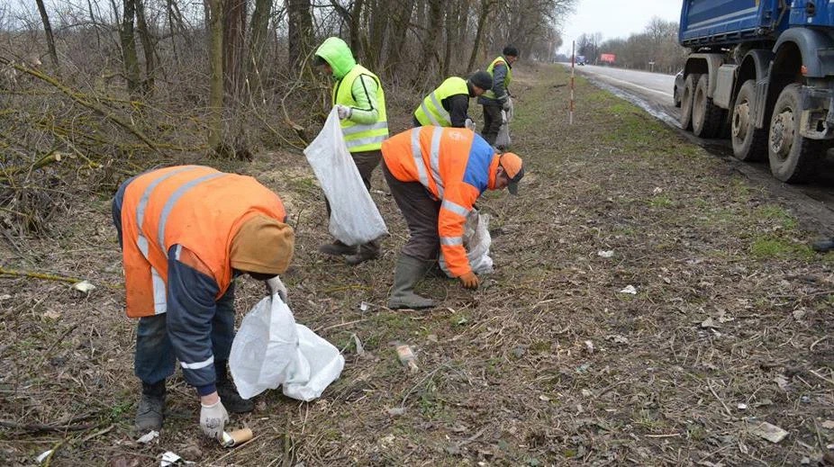 На фотографии работники в светоотражающей одежде собирают мусор на обочине дороги.  Они используют мешки для мусора, а рядом стоит грузовик.