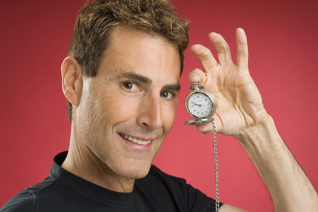 Ури Геллер держит карманные часы на фоне красной стены
